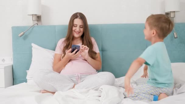 Piccolo figlio fastidioso e pasticciare con la madre sdraiata a letto e utilizzando smartphone. Concetto di bambini allegri e felicità familiare. — Video Stock