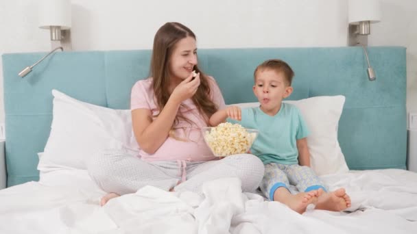 Feliz madre sonriente con su hijo pequeño acostado en la cama y comiendo palomitas de maíz de un tazón grande. Concepto de niños alegres y felicidad familiar. — Vídeo de stock