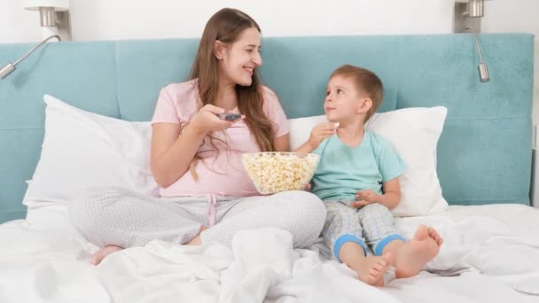 Boldog mosolygós anya kisfiúval, aki popcornt eszik és filmet néz az ágyban reggel. — Stock videók