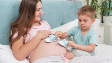 Gülümseyen hamile kadın, küçük oğlunun koca göbeğinde bebek botlarıyla oynamasına bakıyor. Neşeli çocuklar ve bebek bekleyen aile mutluluğu kavramı