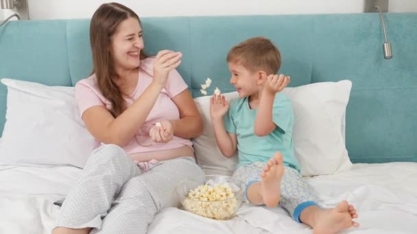 SLow pohyb šťastné laughign rodiny v pyžamu mají boj s popcornem při sledování karikatury v posteli. Koncept veselých dětí a rodinného štěstí.