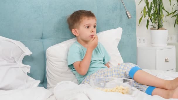 Menino de pijama deitado na cama e comendo pipocas enquanto assiste TV de manhã. — Vídeo de Stock