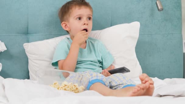 Mały chłopiec w piżamie je popcorn w łóżku rano i włącza kreskówki w TV z pilotem — Wideo stockowe