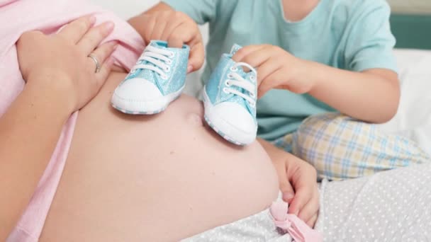 Een close-up van een jongetje dat babyschoentjes speelt op zijn zwangere moeder. Concept van vrolijke kinderen en familie geluk verwacht baby — Stockvideo