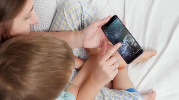 Närbild av ung kvinna som visar baby ultraljud bild till sin lille son på smartphone. Begreppet sjukvård och familj lycka väntar barn — Stockvideo