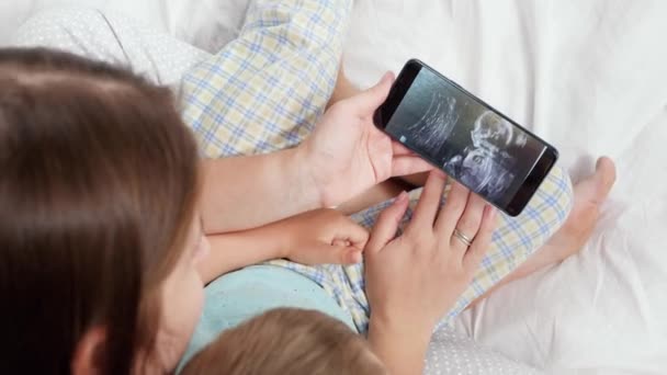 Mladá matka a malý syn v pyžamu leží v posteli a dívají se na ultrazvukový obraz budoucího dítěte a bratra. Koncepce zdravotní péče a rodinné štěstí čeká dítě — Stock video