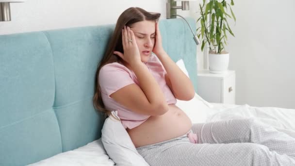 Een jonge zwangere vrouw in een pyjama die hoofdpijn heeft en haar hoofd raakt. Concept van gezondheidszorg en pijn tijdens de zwangerschap — Stockvideo