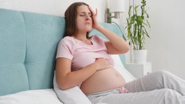 Jeune femme enceinte se sentant mal à l'aise souffrant de maux de tête et couchée au lit. Concept de soins de santé et de douleur pendant la grossesse — Video