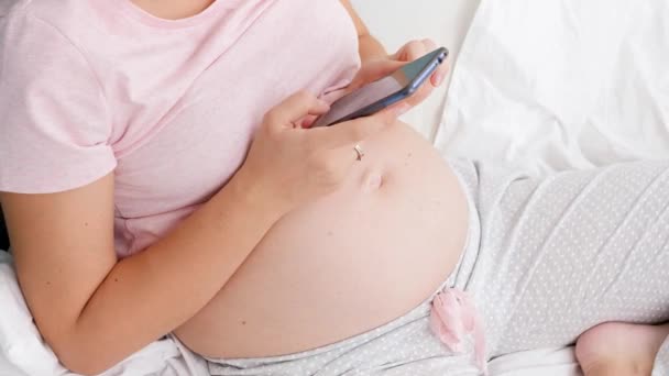Top weergave van jonge zwangere vrouw in pyjama typen bericht op smartphone terwijl zittend op bed in de ochtend — Stockvideo