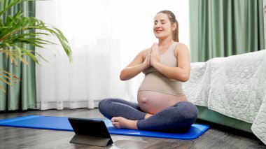 Genç hamile kadın spor minderinde oturuyor ve tablet bilgisayarda yoga yapıyor. Hamilelik boyunca sağlıklı yaşam tarzı, sağlık ve spor kavramı