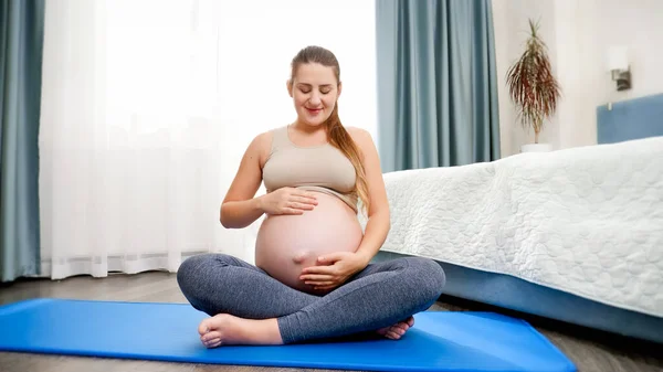 Mooie glimlachende zwangere vrouw mediteren op fitness mat bij groot raam en strelen haar dikke buik. Concept van gezonde levensstijl, gezondheidszorg en sport tijdens de zwangerschap — Stockfoto