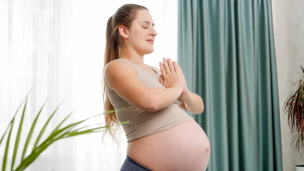 Giovane donna incinta meditando e praticando yoga alla grande finestra contro cielo luminoso. Concetto di stile di vita sano, assistenza sanitaria e sport durante la gravidanza — Foto Stock