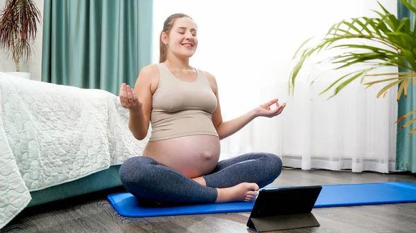 Donna incinta sorridente che utilizza tablet e pratica yoga sul tappeto fitness a casa. Concetto di stile di vita sano, assistenza sanitaria e sport durante la gravidanza — Foto Stock