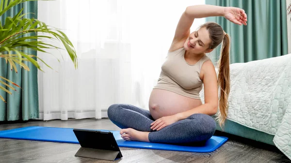 İnternette yoga yapan gülümseyen hamile kadın tablet bilgisayarına bakıyor. Hamilelik boyunca sağlıklı yaşam tarzı, sağlık ve spor kavramı — Stok fotoğraf