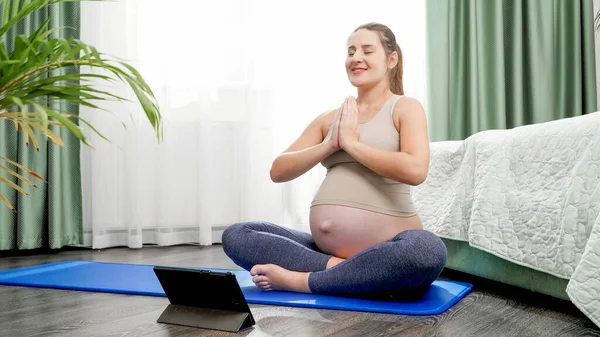 Ung gravid kvinna sitter på träningsmatta och utövar yoga online på surfplatta dator. Begreppet hälsosam livsstil, hälsovård och idrott under graviditeten — Stockfoto