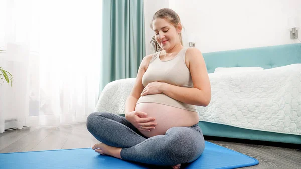 Bella donna incinta sorridente in abito sportivo seduta sul tappeto fitness e accarezzando la sua grande pancia. Concetto di stile di vita sano, assistenza sanitaria e sport durante la gravidanza — Foto Stock