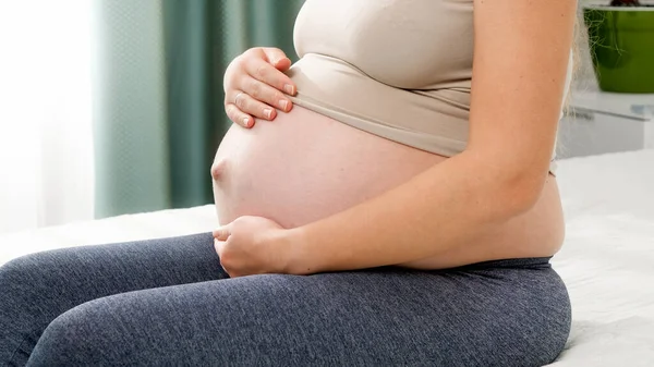 Mulher grávida bonita sentada ao lado da cama e acariciando sua barriga de pântano contra a grande janela. Conceito de estilo de vida saudável, cuidados de saúde e esportes durante a gravidez — Fotografia de Stock