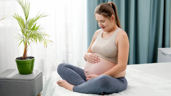 Sonriente mujer embarazada relajada acariciando y tocando su vientre contra una gran ventana en el dormitorio. Concepto de estilo de vida saludable, atención médica y deportes durante el embarazo —  Fotos de Stock