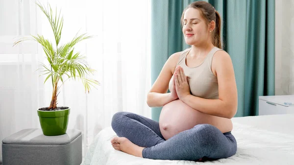 Gyönyörű mosolygó terhes nő mélyen lélegzik, és meditál lótusz ászana az ágyon ellen nagy ablak. Az egészséges életmód, az egészségügy és a sport koncepciója a terhesség alatt — Stock Fotó