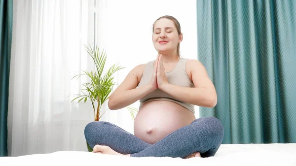 Felice donna incinta sorridente praticare yoga seduto in posizione loto e lloking in macchina fotografica. Concetto di stile di vita sano, assistenza sanitaria e sport durante la gravidanza — Foto Stock