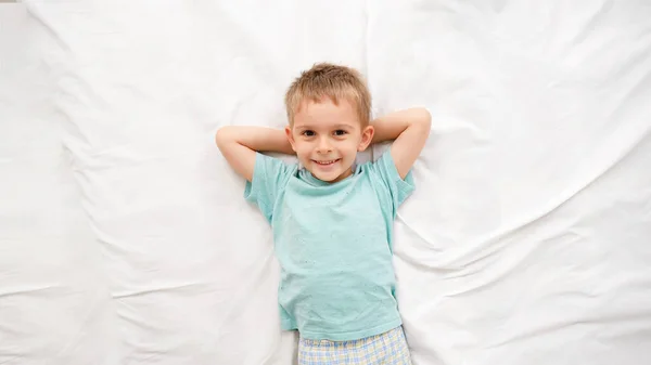 Retrato de vista superior do menino sorridente deitado em lençóis brancos na cama e olhando para a câmera. Conceito de crianças felizes se divertindo em casa — Fotografia de Stock