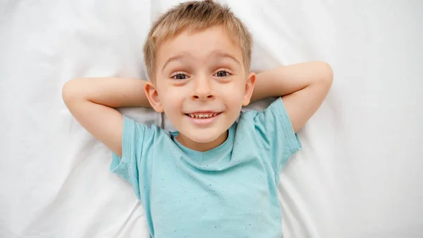 Retrato de un niño sonriente en pijama acostado en una cama grande con sábanas blancas y mirando a la cámara. Concepto de niños felices pasándolo bien en casa — Foto de Stock