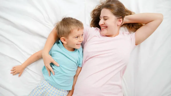 Visão superior da alegre jovem mãe feliz com o pequeno filho em pijama deitado na cama e sorrindo. COncept de parentalidade e felicidade familiar — Fotografia de Stock