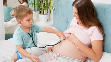 Doktorda oynayan sevimli çocuk ve steteskopla hamile annenin kalp atışlarını dinliyor. Hamilelik süresince sağlık ve tıbbi muayene kavramı