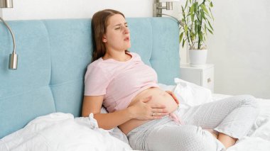 Genç hamile bir kadın karnında ağrı hissediyor yatakta dinleniyor ve karnını okşuyor. Hamilelik boyunca sağlık ve alkol kullanımı