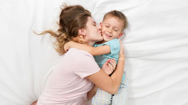 Top view portrait of little boy in pajamas hugging and embracing his smiling mother lying with him on bed with white sheets. Concept of parenting, loving children and family happiness — Stock Photo, Image