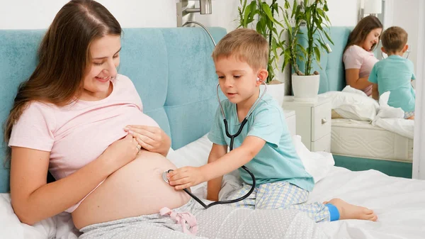Niño pequeño escuchando madres embarazadas vientre grande con estetoscopio. Concepto de salud y examen médico durante el embarazo — Foto de Stock