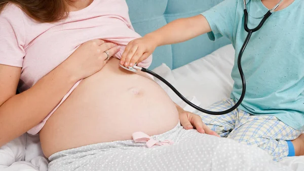 妊娠中の母親と一緒に聴診器を演奏し、彼女のハートビートを聞いている小さな男の子。妊娠中の健康診断の概念 — ストック写真