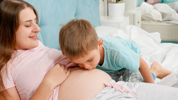 Cute adorable boy kissing and listening to big belly of his pregnant mother lying on bed. Concept of loving children and family happiness expecting baby — Stock Photo, Image