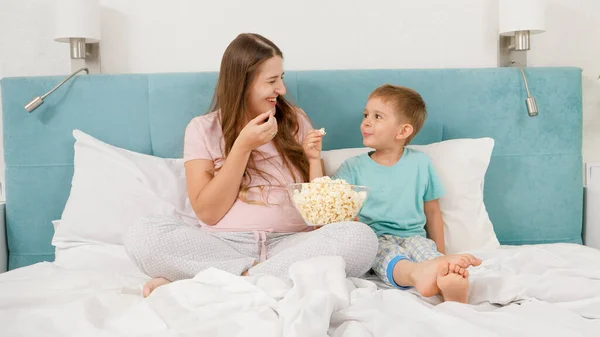 Mãe sorridente feliz com o filho da criança deitado na cama e comendo pipocas de tigela grande. Conceito de crianças alegres e felicidade familiar. — Fotografia de Stock