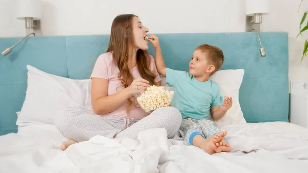 Petit garçon tout-petit avec sa jeune mère en pyjama couchée au lit le week-end et mangeant du pop-corn dans un grand bol. Concept d'enfants gais et de bonheur familial. — Photo
