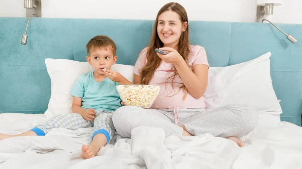Lindo niño pequeño con madre usando pijamas viendo dibujos animados en la mañana del fin de semana y comiendo palomitas de maíz en la cama. Concepto de niños alegres y felicidad familiar. — Foto de Stock