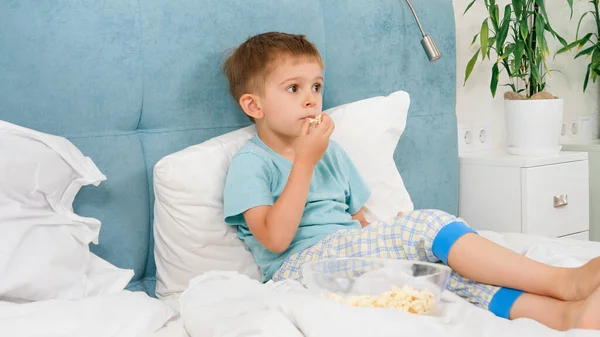 Маленький мальчик в пижаме лежит в постели и ест попкорн во время просмотра телевизора по утрам. — стоковое фото