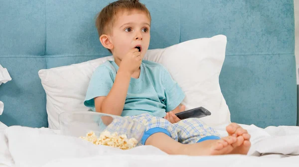 Menino de pijama comendo pipocas na cama de manhã e trocando desenhos animados na TV com controle remoto — Fotografia de Stock