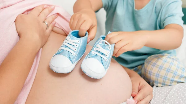 Närbild av liten pojke spelar med bebis skor på sin gravida mamma stor mage. Begreppet glada barn och familj lycka väntar barn — Stockfoto