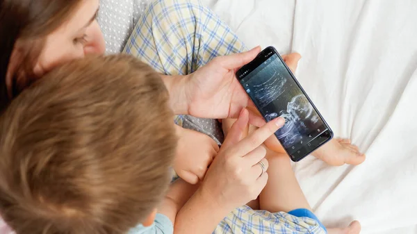 Primer plano del niño pequeño mirando la imagen de ultrasonido de su hermano bebé nonato en el teléfono inteligente de las madres. Concepto de salud y felicidad familiar esperando bebé —  Fotos de Stock