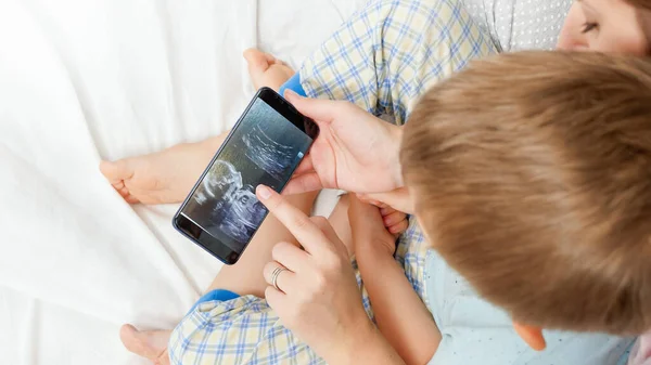 Vista dall'alto della madre incinta con il figlio maggiore che guarda l'immagine ecografica del suo futuro bambino. Concetto di assistenza sanitaria e felicità familiare in attesa del bambino — Foto Stock