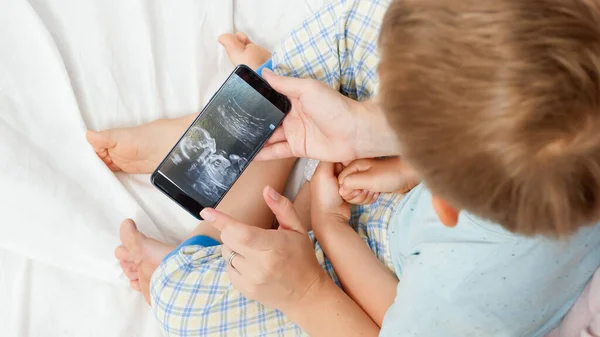 Primer plano de la mujer joven que muestra la imagen de ultrasonido del bebé a su hijo pequeño en el teléfono inteligente. Concepto de salud y felicidad familiar esperando bebé —  Fotos de Stock