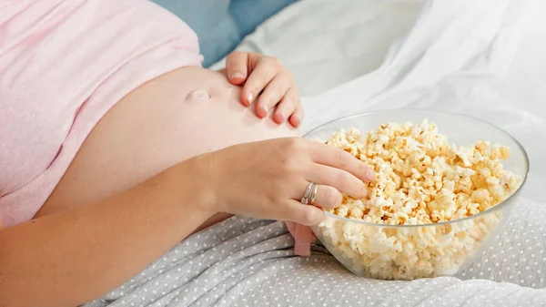 Pijamalı hamile bir kadının TV izlerken yatakta gevşeyip patlamış mısır yemesi. — Stok fotoğraf