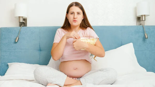 O femeie însărcinată concentrată se uită la film la televizor și mănâncă popcorn din castron mare — Fotografie, imagine de stoc