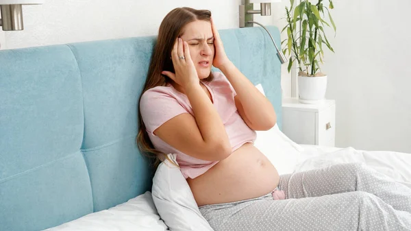 Портрет молодой беременной женщины, страдающей головной болью, лежащей в постели. Концепция здравоохранения и боли во время беременности — стоковое фото