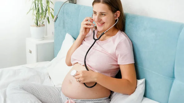 Feliz sorrindo mulher grávida usando estetoscópio para ouvir seu bebê batimento cardíaco. Conceito de cuidados de saúde e exame médico durante a gravidez — Fotografia de Stock