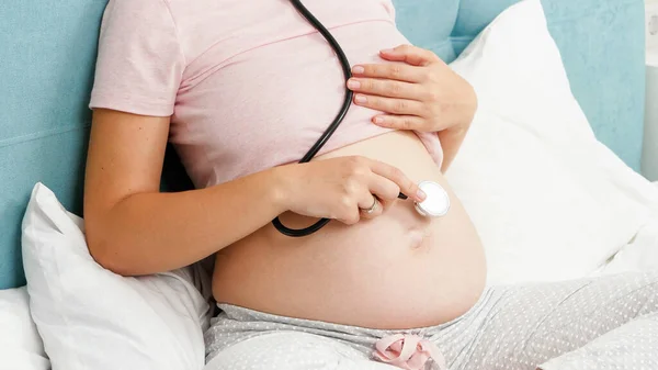 妊娠中の女性の閉鎖は、聴診器を使用して、生まれていない子供と彼女の大きな腹にそれを保持します.妊娠中の健康診断の概念 — ストック写真