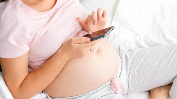 Вид сверху на молодую беременную женщину с большим животом, которая просматривает Интернет и пользуется смартфоном — стоковое фото
