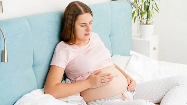 Mujer embarazada joven sintiendo dolor duro en el estómago cogido de la mano en el vientre mientras está acostado en la cama en casa —  Fotos de Stock