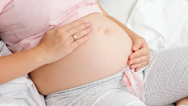 Close-up de jovem grávida acariciando sua barriga grande enquanto se senta na cama. Conceito de parentalidade e expectativa feliz do futuro bebê. — Fotografia de Stock